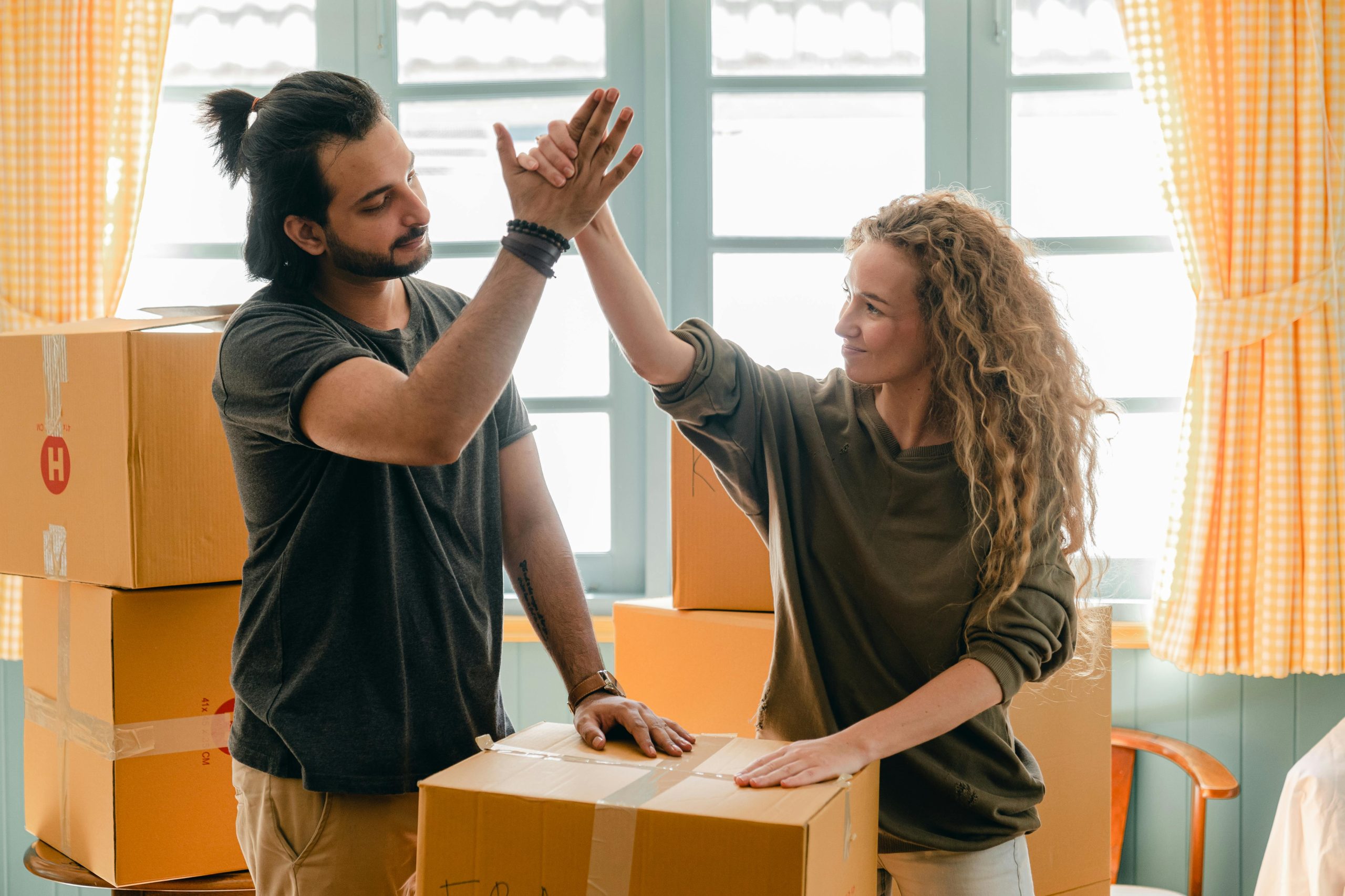 Home Happy woman in casual wear standing near heap of cardboard boxes and giving high five to ethnic boyfriend with ponytail showing agreement while looking at each other